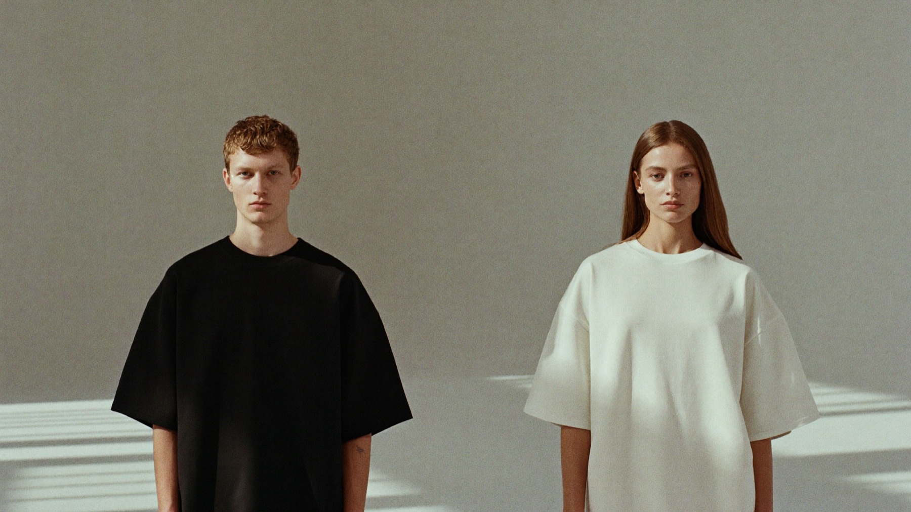 Two models wearing Noir and Écru heavyweight tees in an empty daylight studio